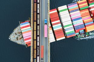 Aerial view of cargo ship sailing under a bridge with trains on railway side and trucks on road side